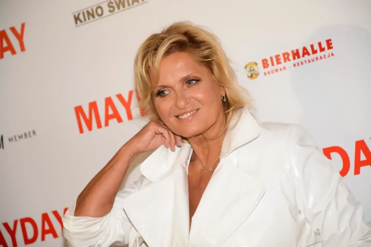 Uśmiechnięta Ewa Kasprzyk w białej, błyszczącej kurtce pozuje na ściance z napisem