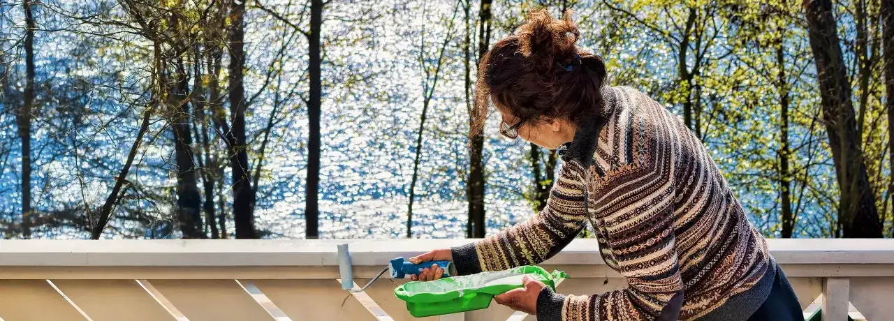 Frau streicht den Balkon wei&szlig;. Sie h&auml;lt eine Farbrolle und eine gr&uuml;ne Farbwanne. Im Hintergrund glitzert Wasser zwischen B&auml;umen.