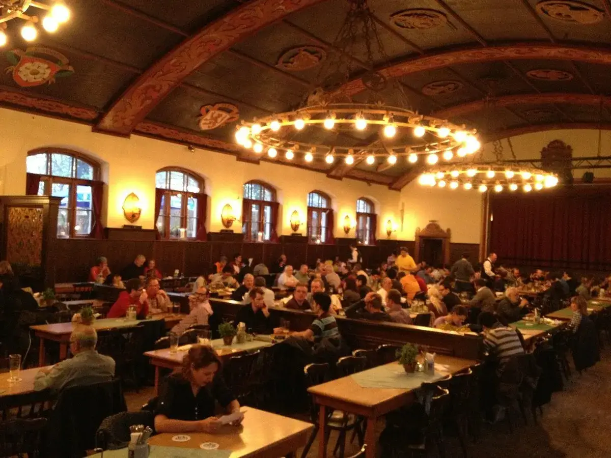Interno di un affollato keller pub con tavoli lunghi, persone che mangiano e bevono, e grandi lampadari accesi.