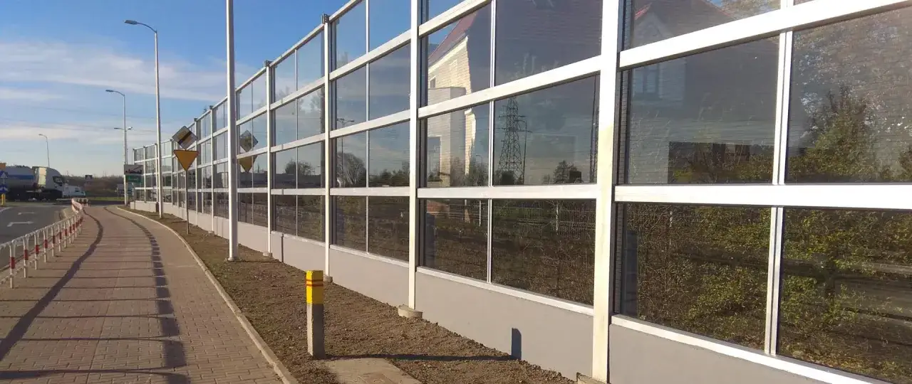 Długie ekrany akustyczne drogowe z przezroczystymi panelami chronią przed hałasem. Widać drogę, chodnik i słupy oświetleniowe.