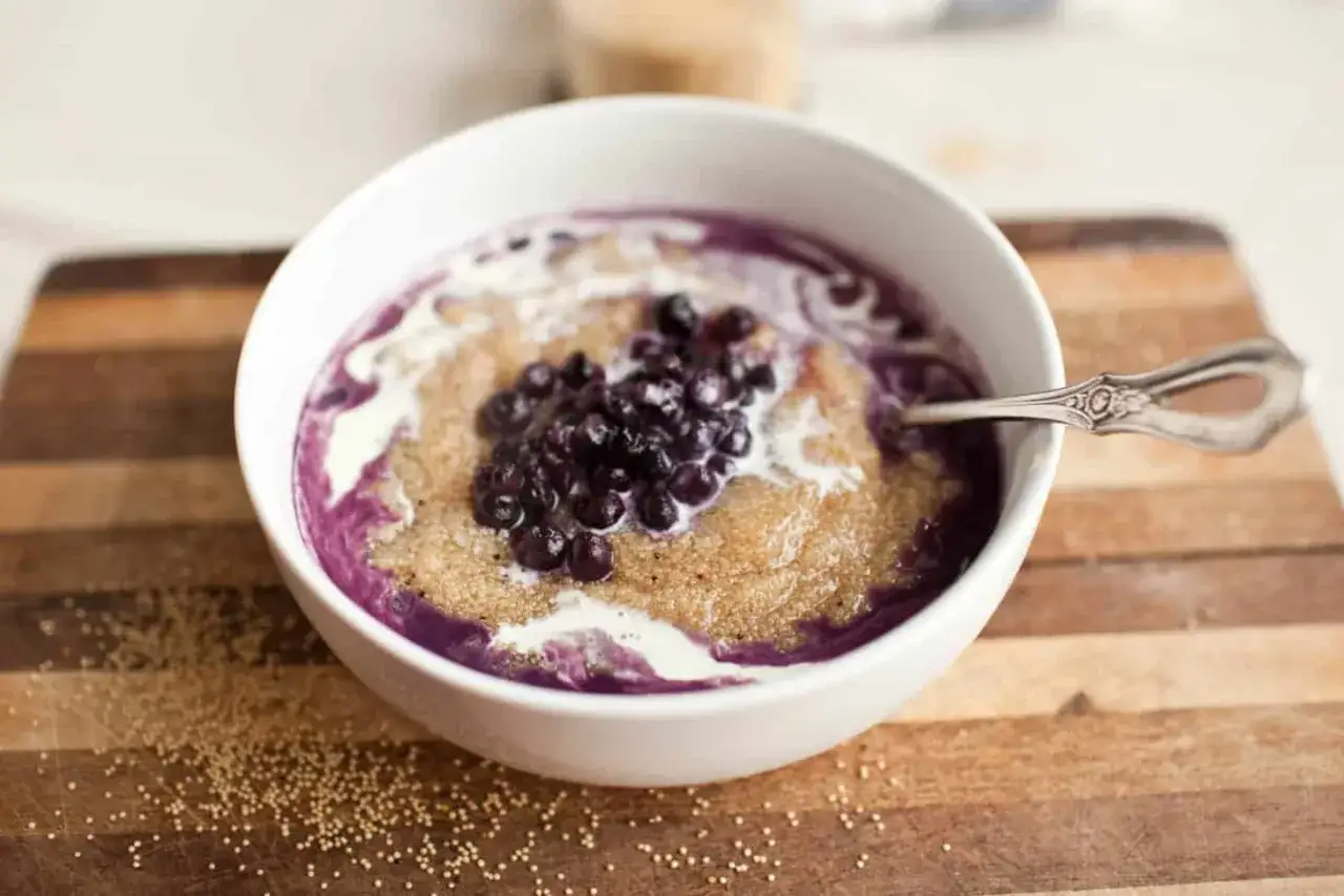 Un taz&oacute;n de amaranto que es un cereal nutritivo, cubierto con ar&aacute;ndanos y crema, servido en una tabla de madera.