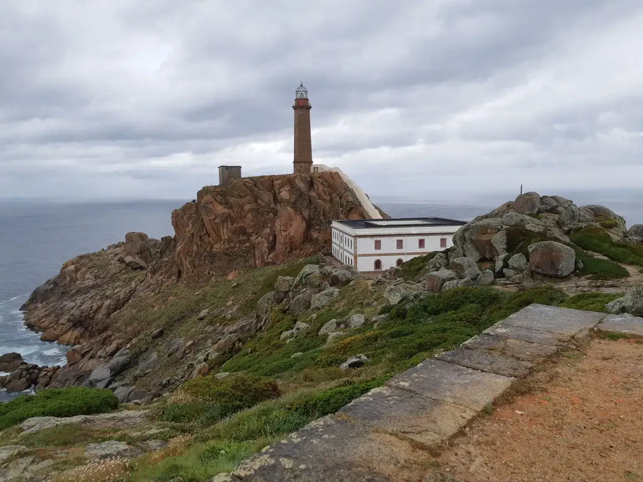 Leuchtturm auf felsiger Klippe an der Costa da Morte, mit wei&szlig;em Geb&auml;ude und st&uuml;rmischem Himmel.