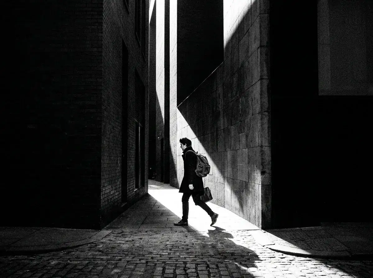 Uomo con zaino cammina in un vicolo stretto, illuminato da un raggio di sole. Una foto di strada drammatica.