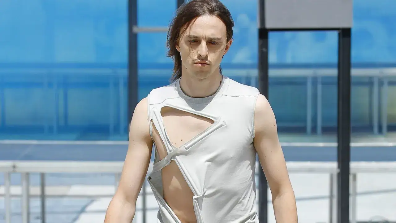 Tommy Cash w szarej, podartej koszulce bez rękaw&oacute;w, prezentuje odważny styl na tle błękitnego budynku.