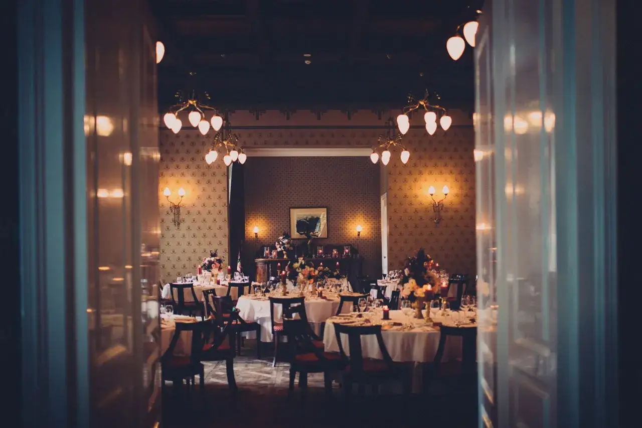 Elegancka sala bankietowa z nakrytymi stołami, ozdobiona kwiatami i świecami. Wnętrze przypomina luksusowe hotele w pałacach, tworząc atmosferę wyjątkowego wydarzenia.