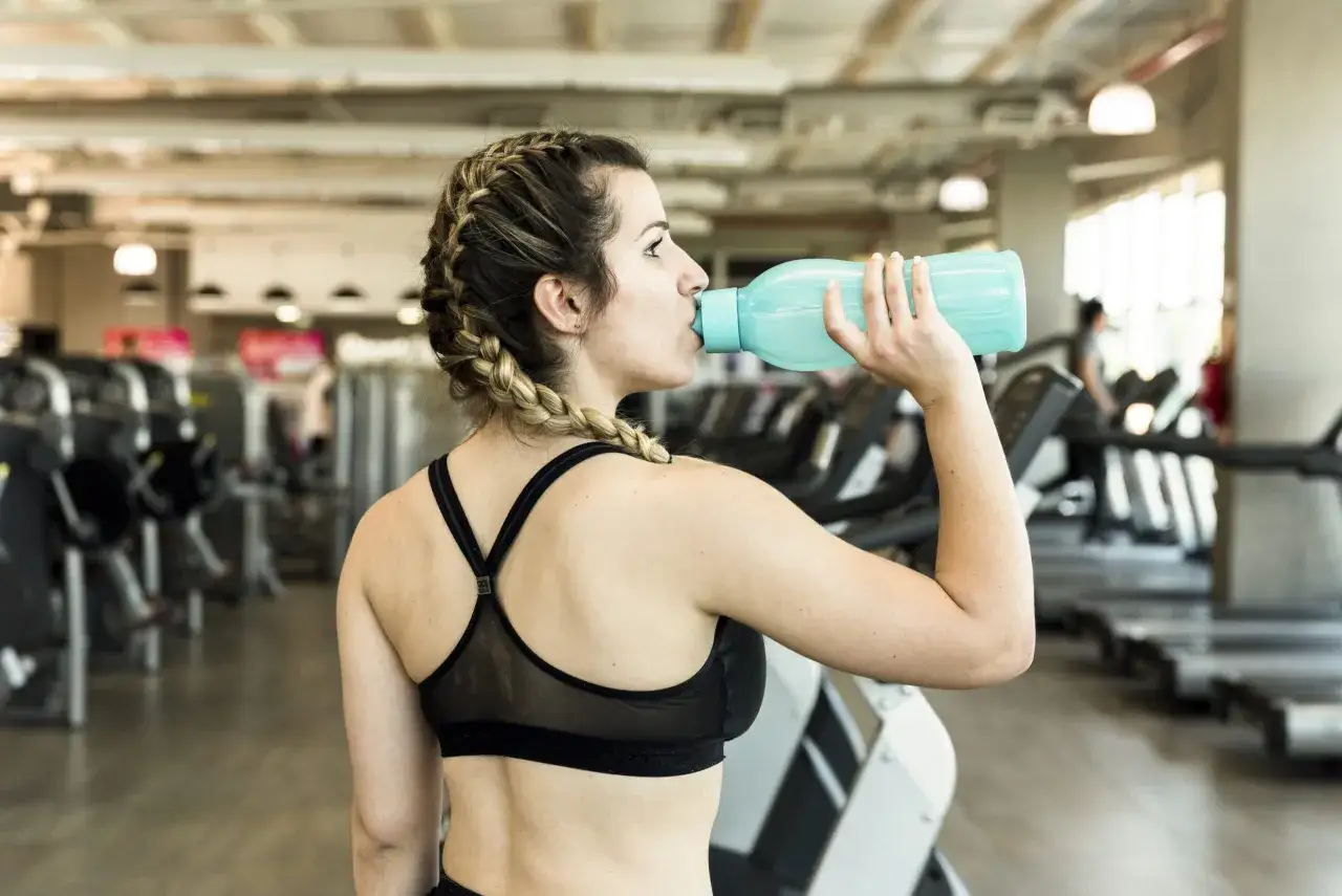 Frau trinkt im Fitnessstudio aus einer blauen Flasche. Sie informiert sich über Kreatin Nebenwirkungen Frauen, um sicher zu trainieren.
