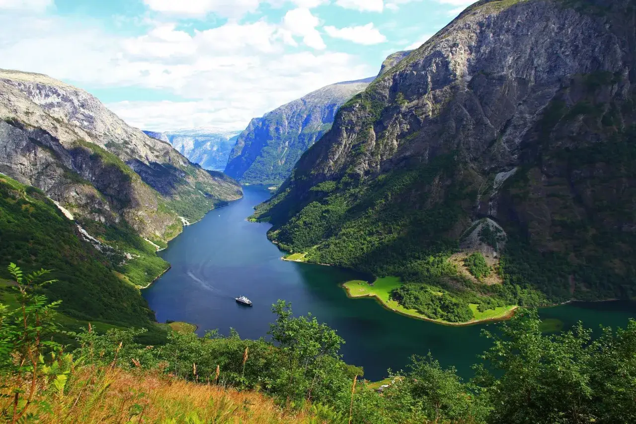 Majestatyczny fiord otoczony zielonymi, skalistymi zboczami. Po wodzie płynie statek, a na brzegu widać małą osadę.