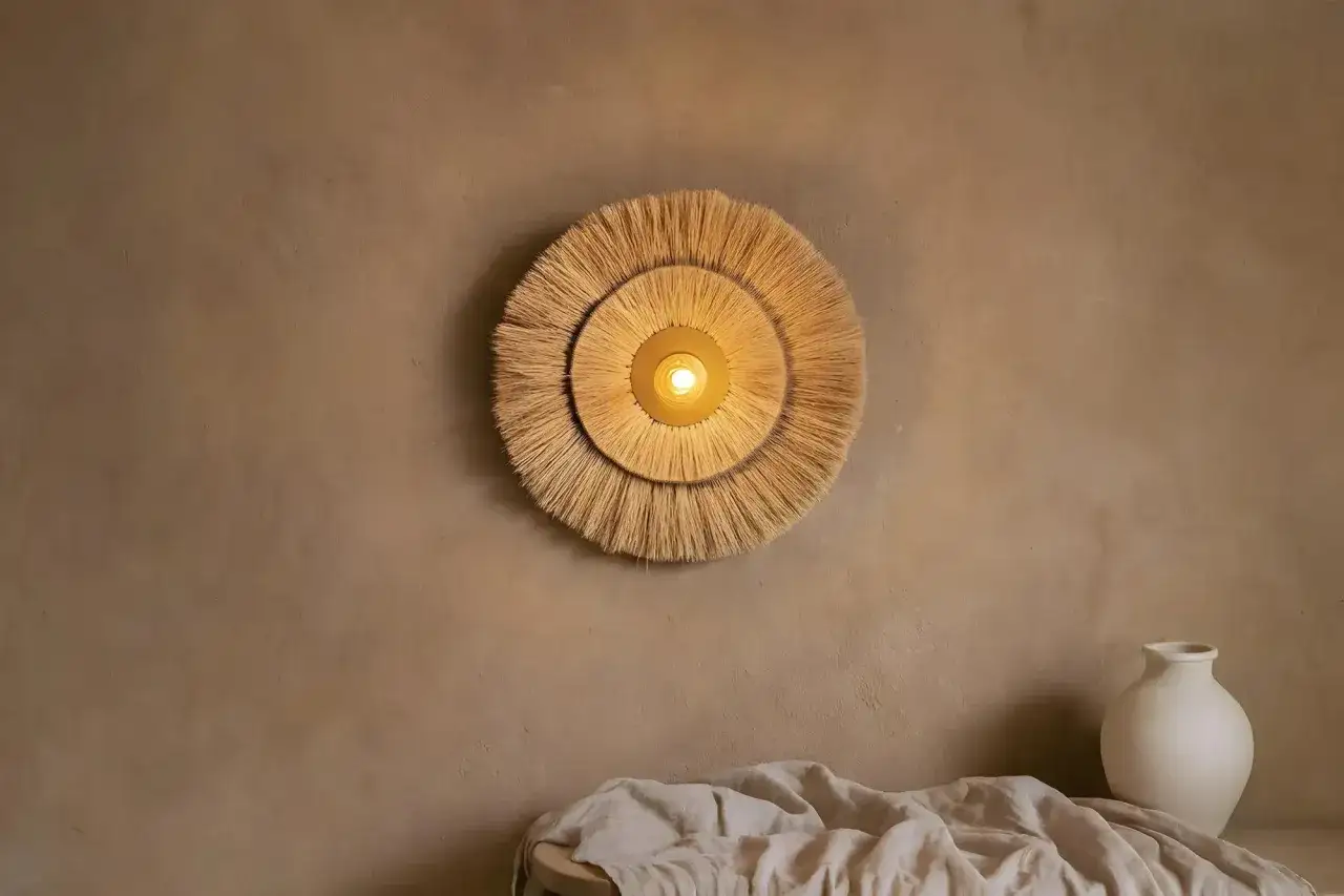 Un applique mural en paille tress&eacute;e, forme soleil, &eacute;claire une pi&egrave;ce aux tons chauds. Un vase blanc et du linge compl&egrave;tent la sc&egrave;ne.