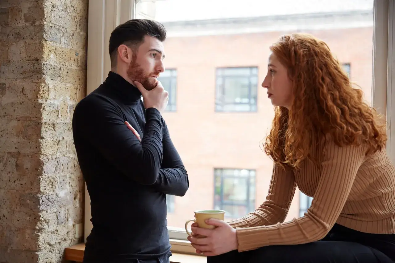 Pareja sentada junto a una ventana, procesar amor en sus miradas. &Eacute;l, pensativo; ella, con una taza.