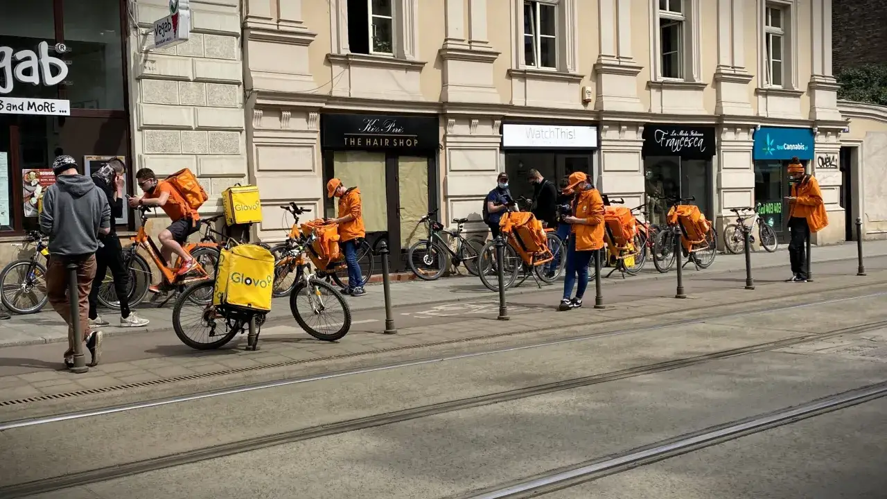 Kurierzy rowerowi z ż&oacute;łtymi torbami Glovo czekają na zlecenia. Ich zarobki zależą od liczby dostaw.