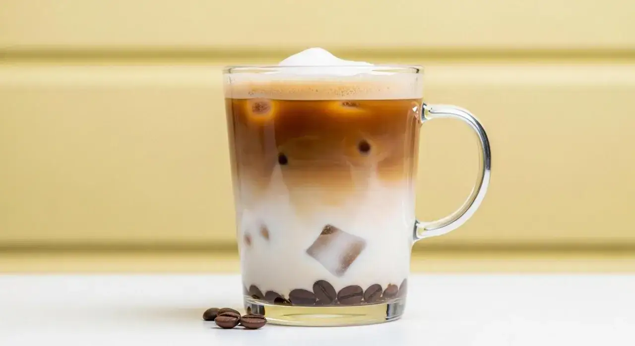 En svalkande cafe frappe med lager av mjölk och kaffe, toppad med skum. Kaffebönor ligger vid glaset.