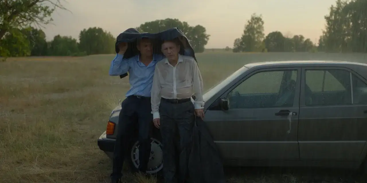 Dwóch mężczyzn w deszczu pod płaszczem obok samochodu. Scena z filmów z Janem Englertem.