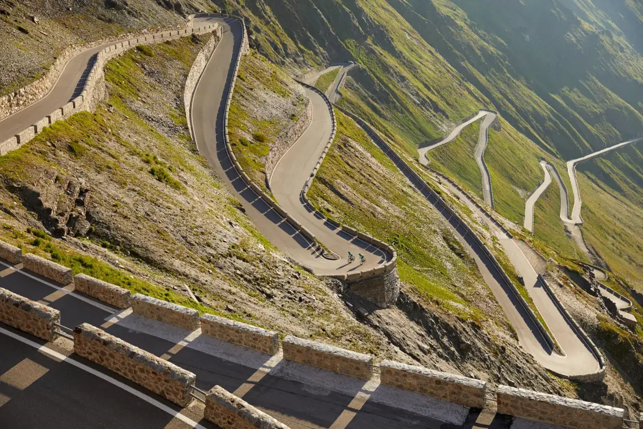 Serpentinen der Stilfser Joch Karte schlängeln sich den Berg hinauf. Zwei Radfahrer genießen die Aussicht.