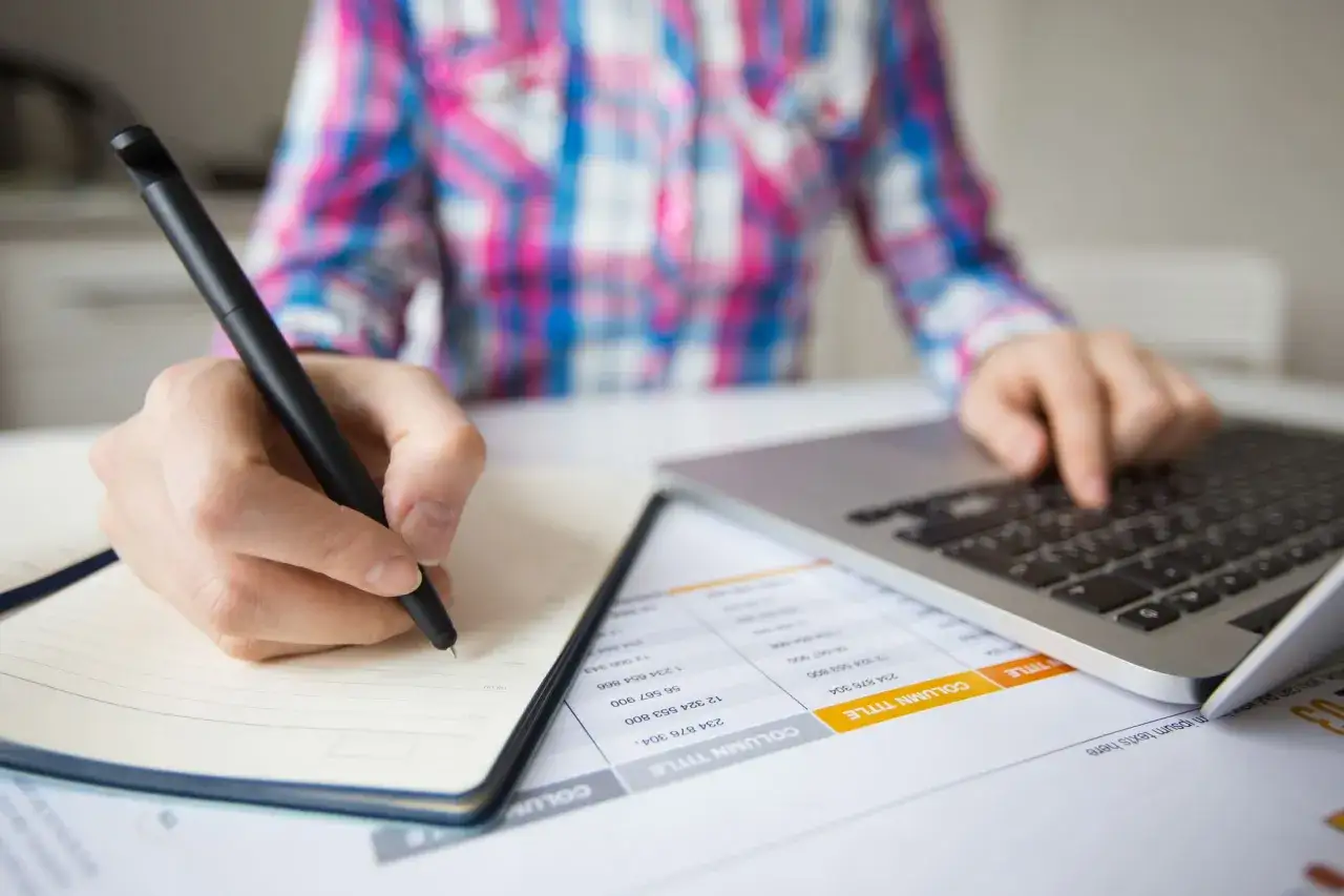 Studente universitario lavoratore scrive appunti su un quaderno mentre usa il laptop.