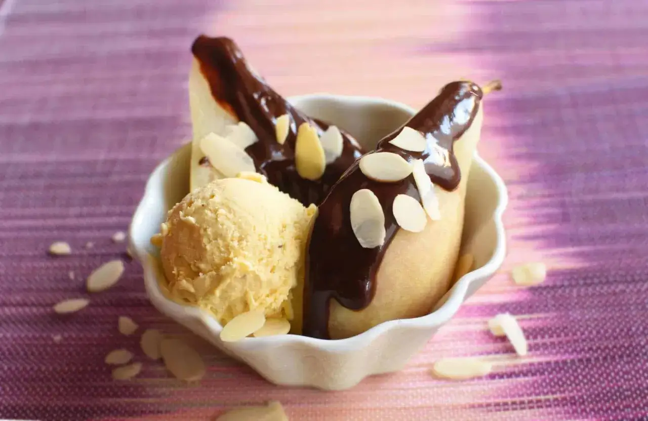 Une poire Belle Hélène, nappée de chocolat chaud et garnie d'amandes effilées, servie avec une boule de glace à la vanille dans un bol blanc.