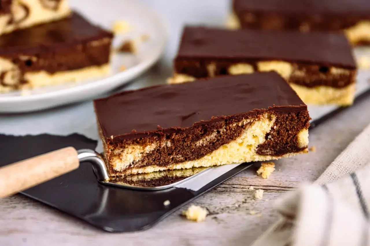 Ein Stück saftiger Marmorkuchen vom Blech, mit Schokoladenglasur, wird auf einem Kuchenheber präsentiert.