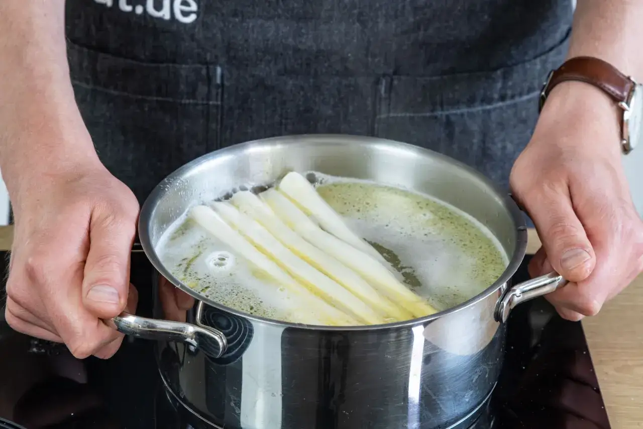 Spargel kochen: Ein Koch hält einen Topf mit siedendem Wasser, in dem weißer Spargel gart. Ob mit oder ohne Deckel, das Ergebnis ist köstlich.