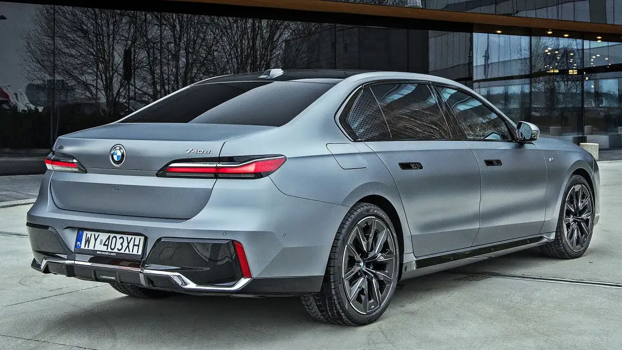 Szare BMW M7 z nowoczesnymi światłami LED i eleganckimi felgami stoi przed budynkiem z lustrzaną fasadą.