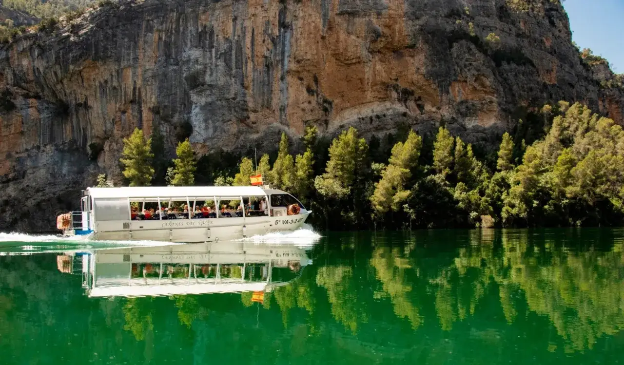 Barco tur&iacute;stico navega por las aguas verdes de los ca&ntilde;ones del J&uacute;car, reflejando la imponente pared rocosa y la vegetaci&oacute;n.