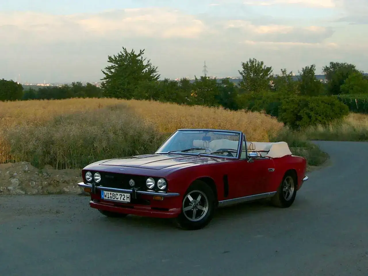 En röd Jensen cabriolet står parkerad vid en lantlig väg, med ett fält av höga gräs i bakgrunden.
