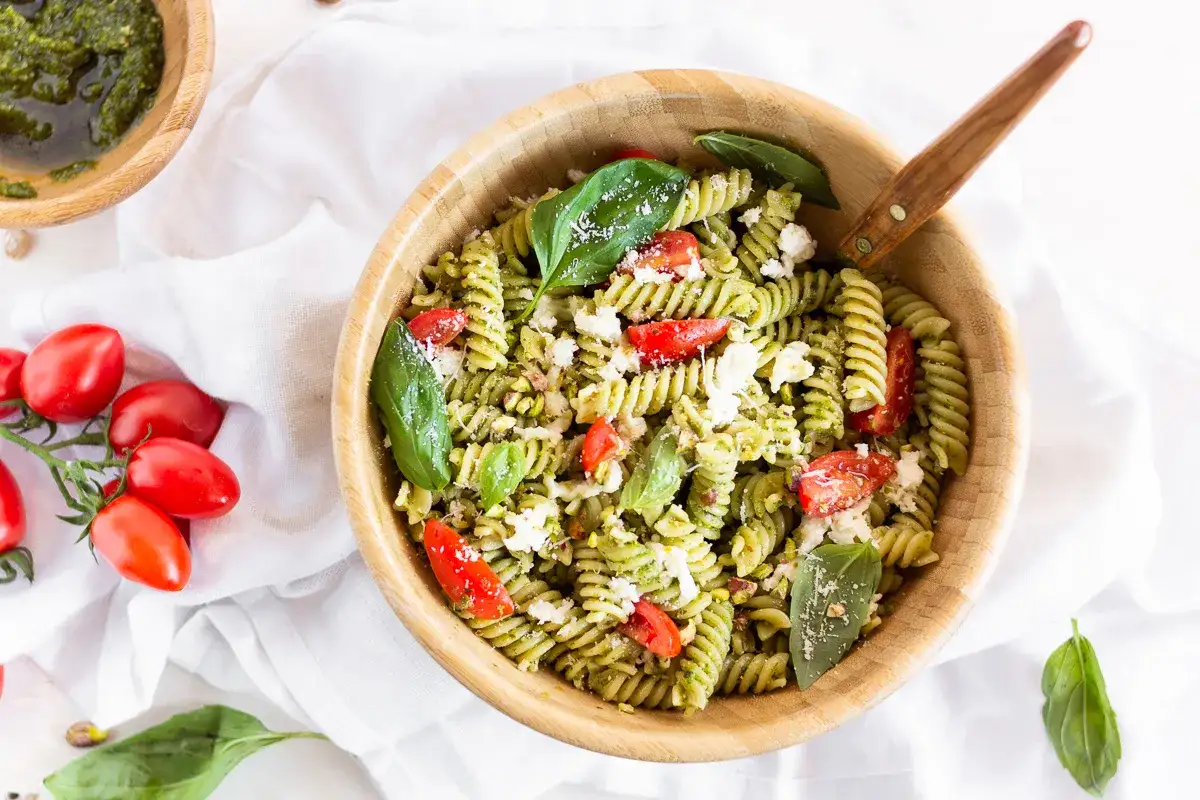 Un'insalata di pasta fredda al pesto, con pomodorini, feta e basilico, servita in una ciotola di legno.