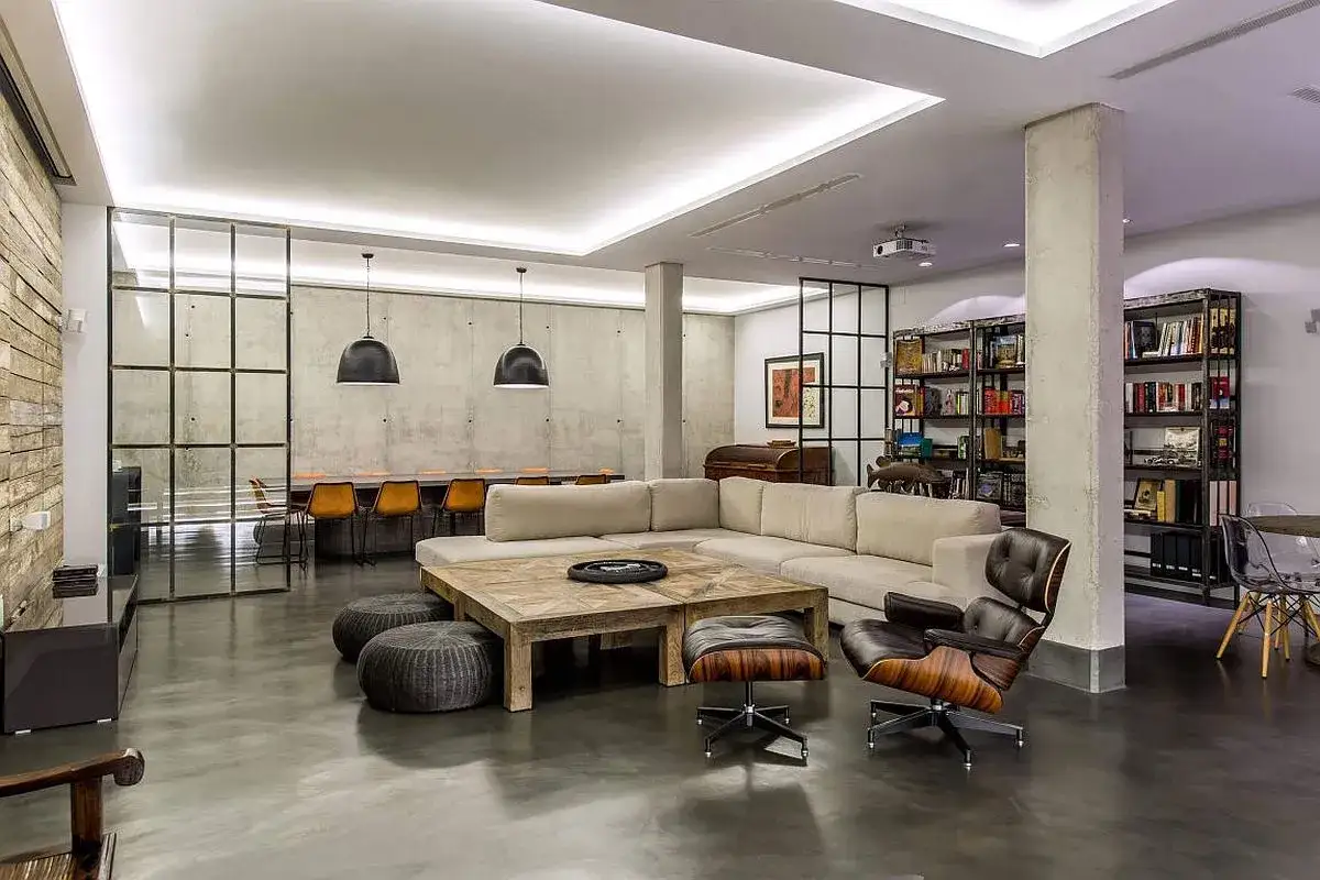 Ger&auml;umiges Wohnzimmer im industrial style mit Betonw&auml;nden, gro&szlig;en Fenstern, bequemen Sofas und einem Holztisch.