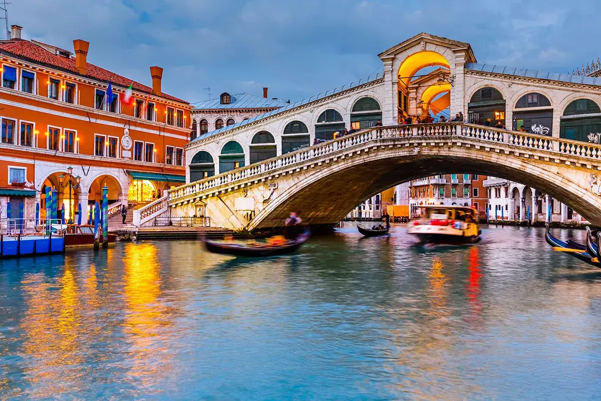 Il Ponte di Rialto a Venezia, con gondole e vaporetti che navigano sul Canal Grande, illuminato al tramonto.