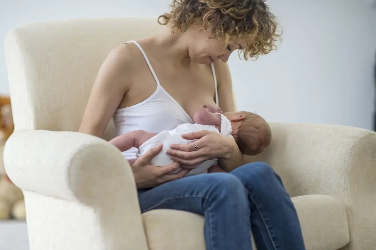 Une m&egrave;re aux cheveux boucl&eacute;s allaite son b&eacute;b&eacute; dans un fauteuil confortable. Un moment de tendresse, loin des soucis du traitement mastite.