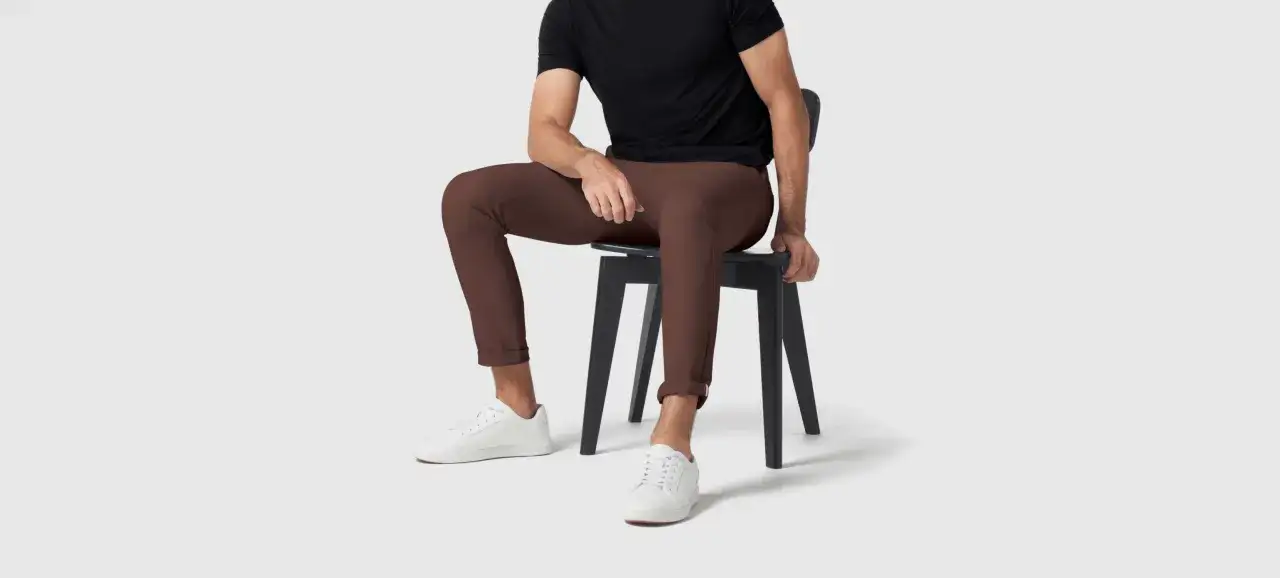 A man in a black shirt and brown pants sits on a black chair, wearing white sneakers.
