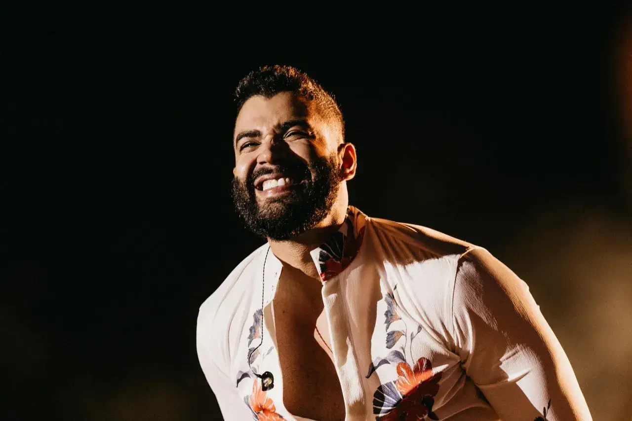 Cantante brasileño famoso joven, con barba y camisa floreada, ríe en el escenario.