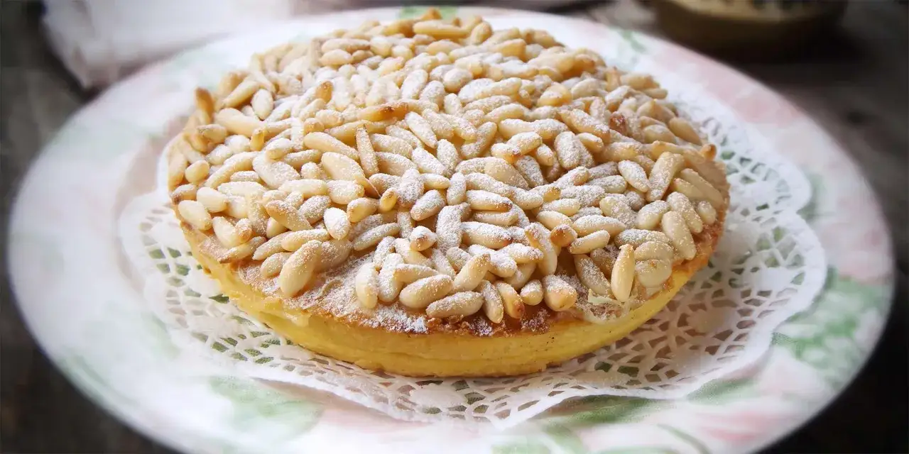 Tarta de piñones, un postre dorado cubierto de piñones tostados y espolvoreado con azúcar glas, servido en un plato delicado.
