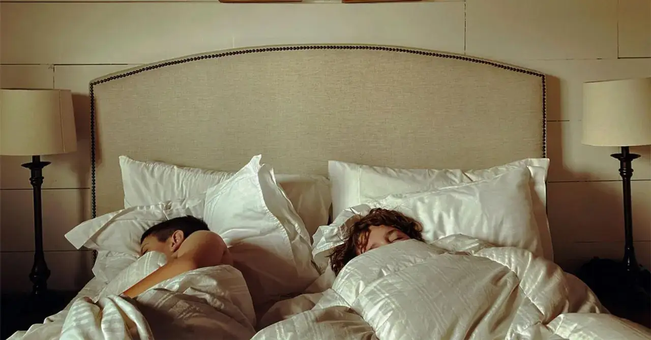 Twee personen liggen vredig samen in bed, omringd door witte lakens en kussens.