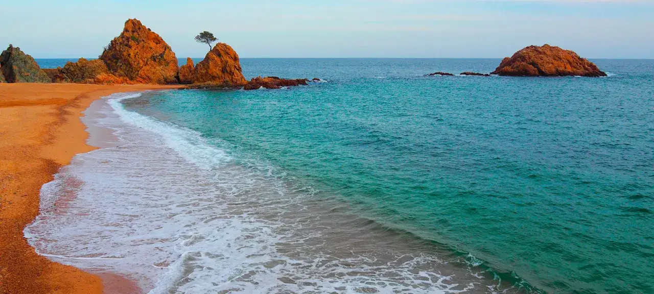 La platja mar menuda, con sus rocas anaranjadas y aguas turquesas, invita a la calma. Una ola rompe suavemente en la arena dorada.