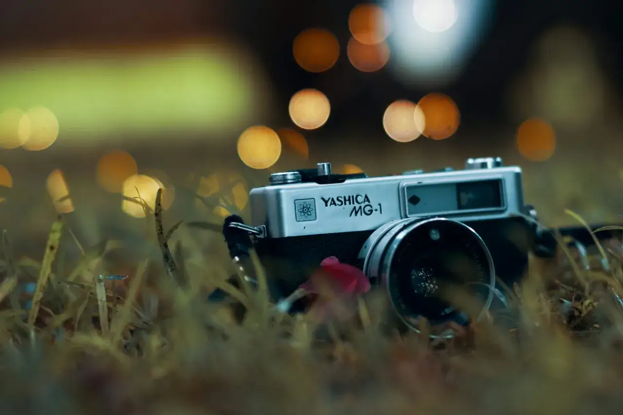A vintage Yashica MG-1 camera rests in the grass, its silver body glinting amidst soft, bokeh lights.