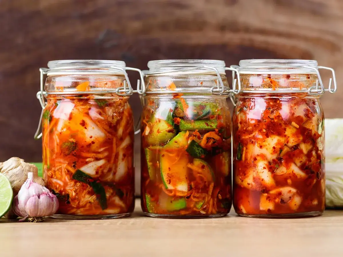 Drei Gl&auml;ser mit selbstgemachtem Kimchi, gef&uuml;llt mit fermentiertem Kohl und Gew&uuml;rzen. Daneben liegen Ingwer, Knoblauch und Limette.