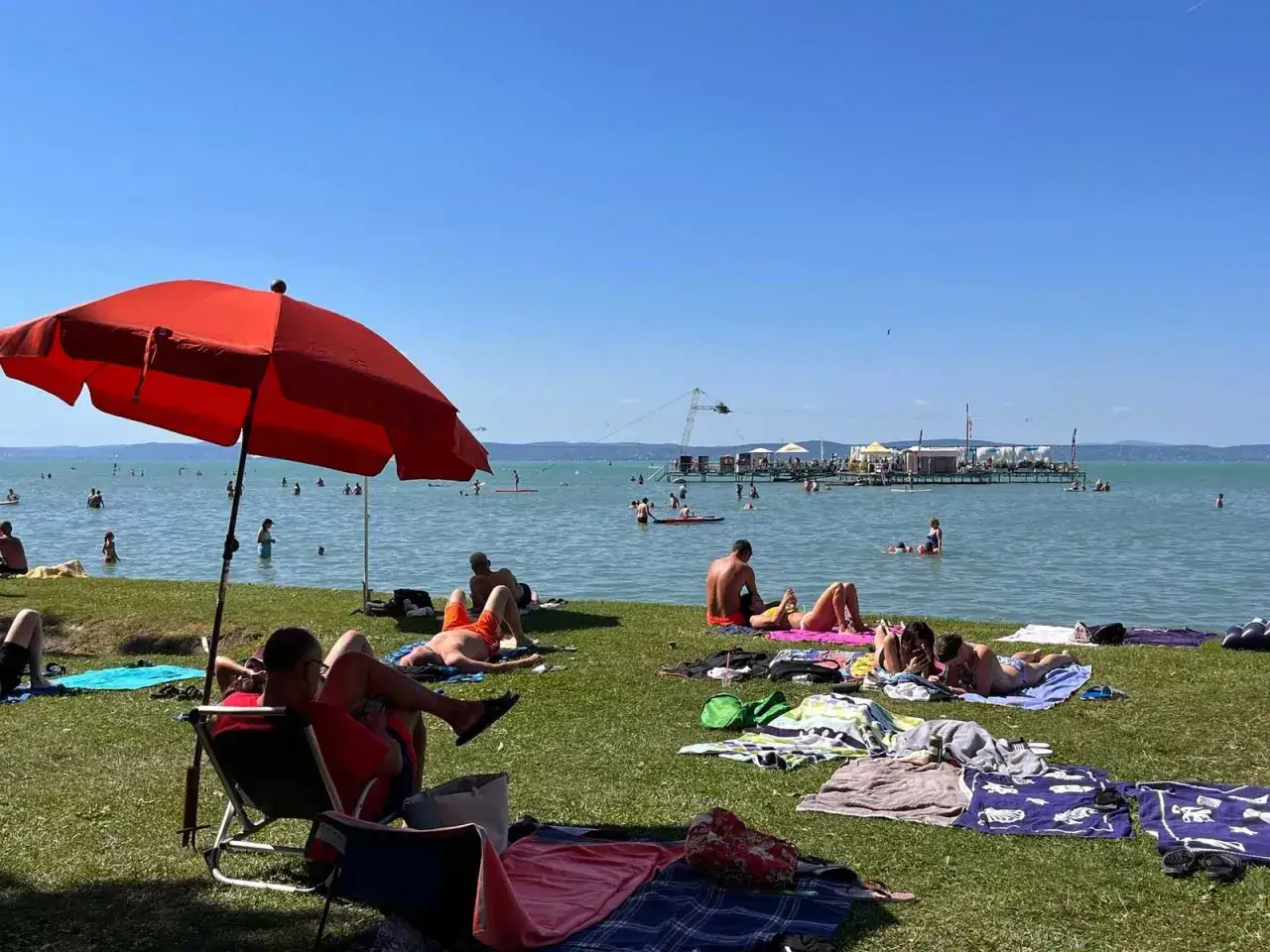 Ludzie odpoczywają na trawie i w wodzie, ciesząc się słońcem i wakacjami na aranypart camping. W tle widać pomost i wyciąg do nart wodnych.
