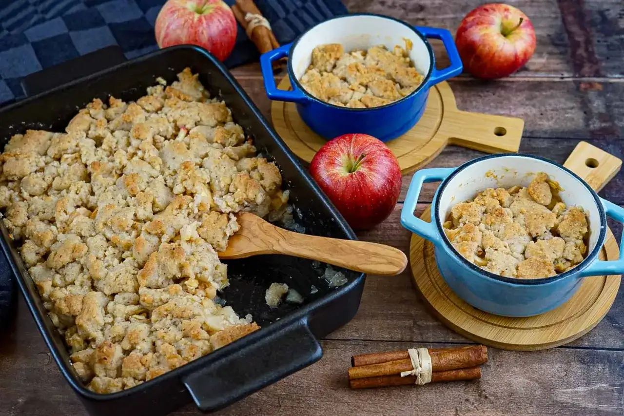 Ein frisch gebackener Apfel Crumble in einer schwarzen Auflaufform und zwei blauen F&ouml;rmchen. Daneben liegen &Auml;pfel und Zimtstangen.