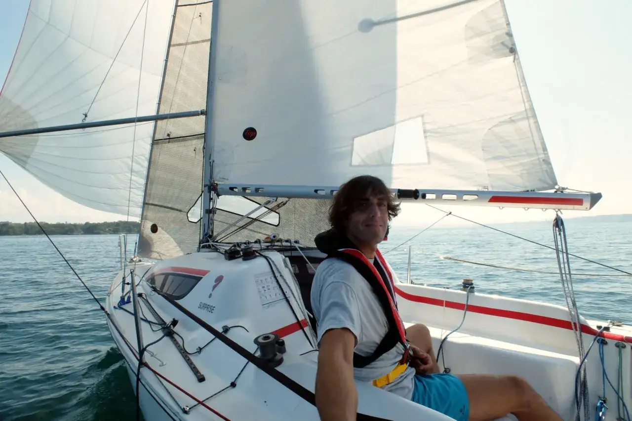 Uomo sorridente su una barca a vela, con le vele spiegate. Un'esperienza di solo vela in una giornata di sole.