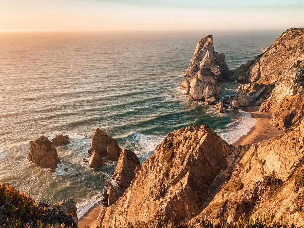 Sonnenuntergang an der Praia da Ursa: dramatische Felsformationen ragen aus dem Meer, eine kleine Bucht mit Sand und Wellen.