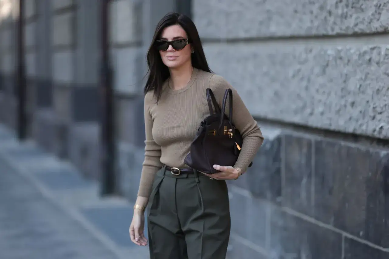 Stylowa it girl w beżowym swetrze i oliwkowych spodniach, z ciemnobrązową torebką i okularami przeciwsłonecznymi.