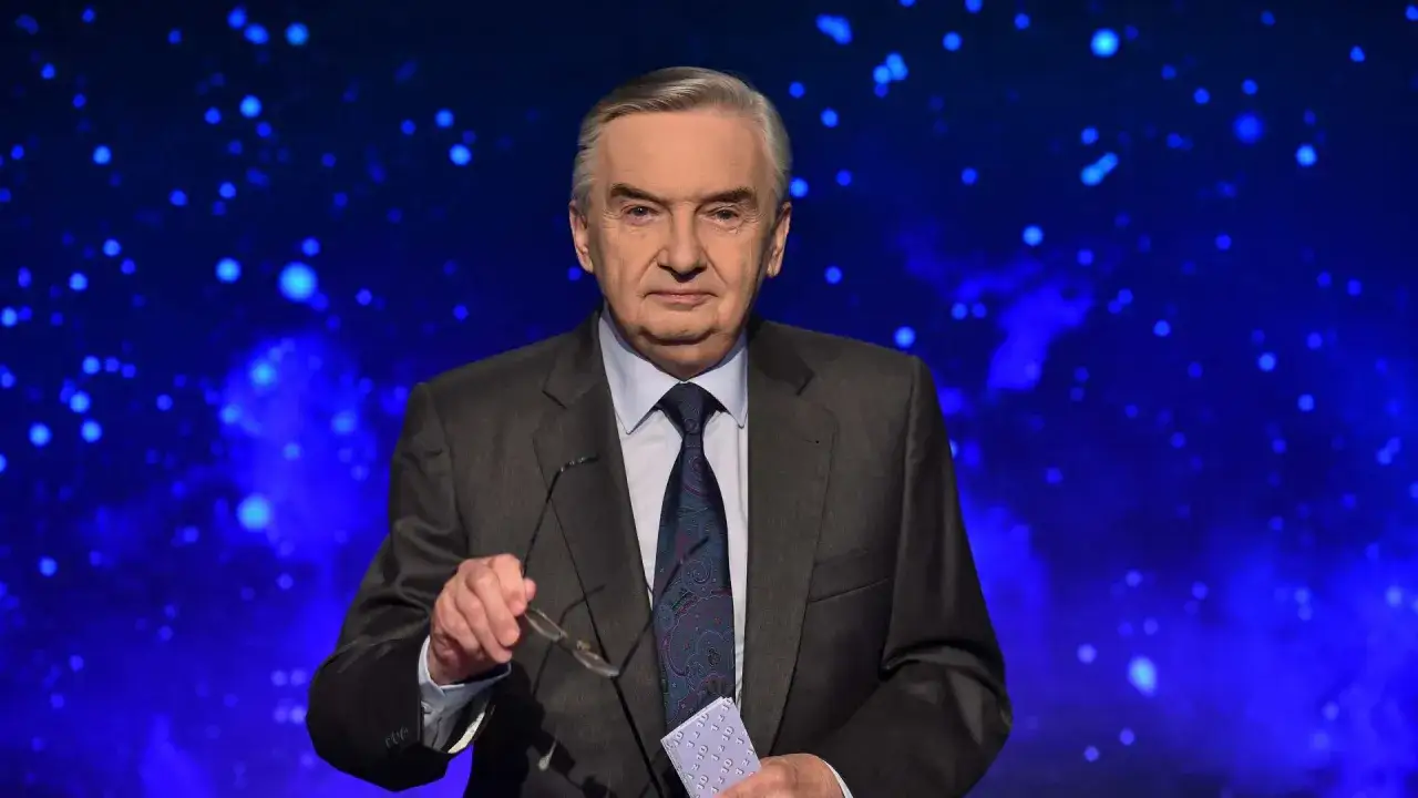 Starsza elegancka osoba w garniturze, trzymająca okulary i kartkę z napisem
