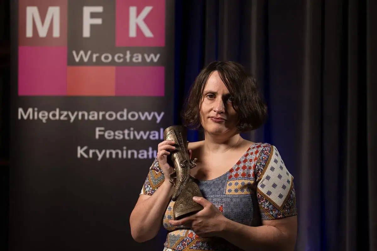 Kobieta trzyma nagrodę wielkiego kalibru na tle banera Międzynarodowego Festiwalu Kryminału we Wrocławiu.