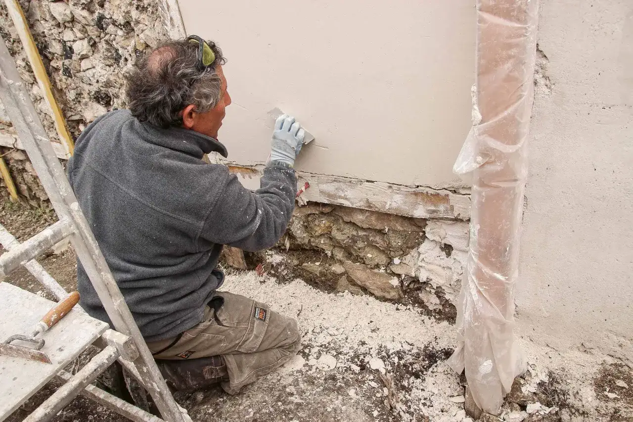 Un ouvrier, agenouillé, applique de l'enduit sur un mur dégradé. Il est en train d'essuyer les plâtres pour une finition parfaite.