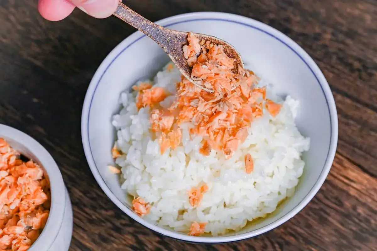 A spoon adds flaked salmon to a bowl of white rice, a simple yet delicious salmon flakes recipe.