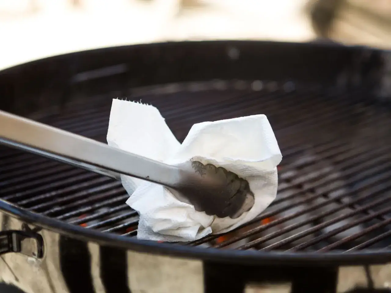 Une pince à barbecue tient un papier absorbant plié, prêt à huiler la grille. La question est : faut il huiler la grille du barbecue ?