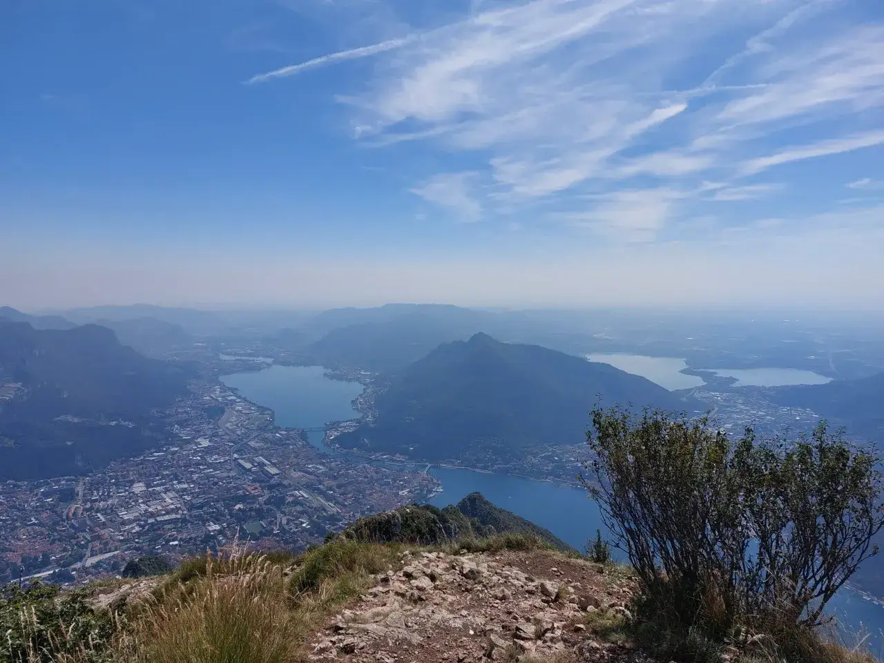 Vista panoramica dal monte Coltignone su laghi e città, con cielo azzurro e nuvole sottili.