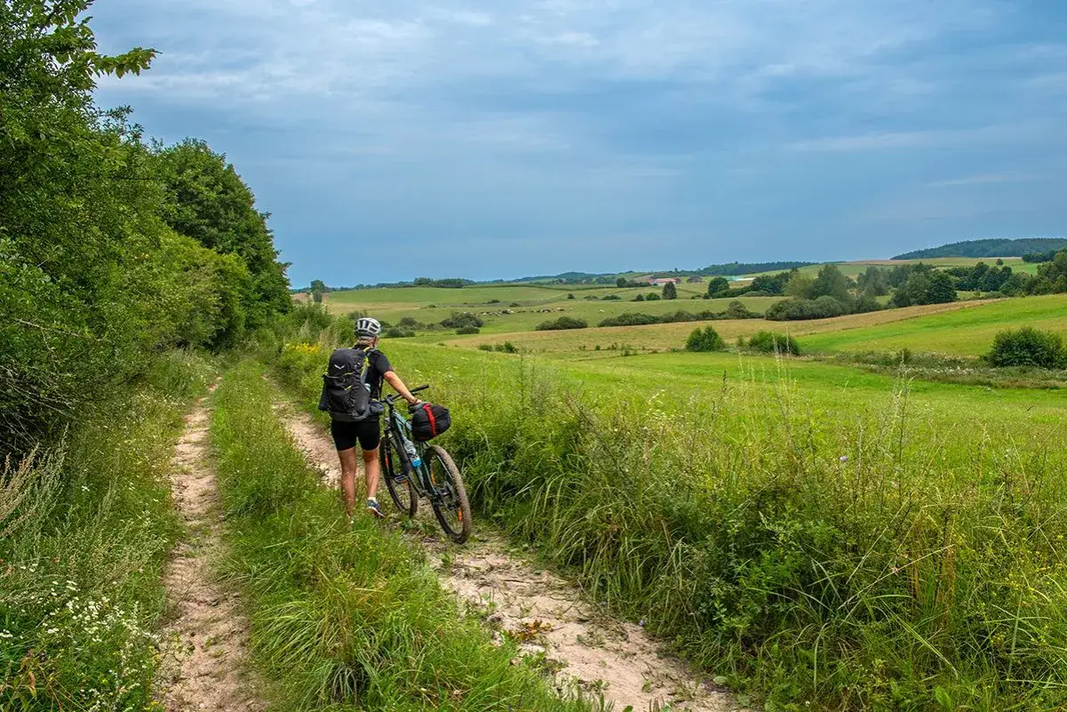 Kobieta z rowerem na szutrowej drodze, odkrywa malownicze **trasy rowerowe Mazury**. Zielone pola i pagórki tworzą sielski krajobraz.
