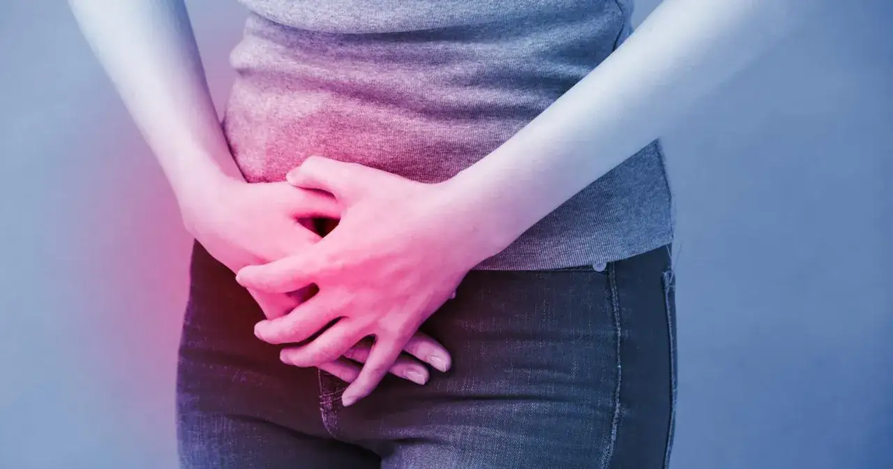 Dłońmi obejmuje podbrzusze, sugerując ból lub dyskomfort, który może wymagać diagnostyki, np. cystoskopii.
