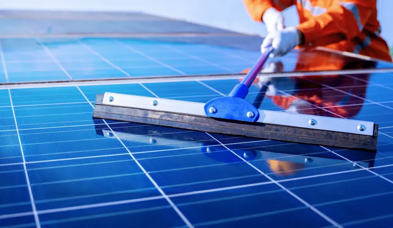Persona con guantes y ropa naranja realiza limpieza paneles solares con rasqueta.