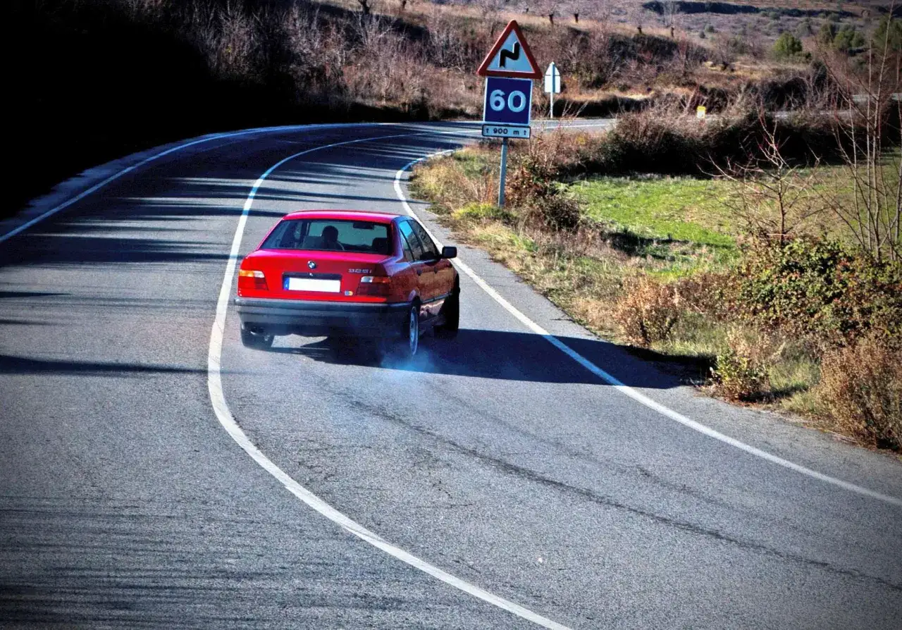 Coche rojo derrapando en curva, dejando humo azul. Señal de curva peligrosa y límite de 60. Se aprecia el **subviraje y sobreviraje** en la maniobra.